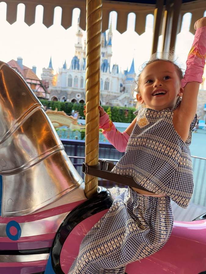 Leonie laughing on a carousel horse with a castle behind her, one arm raised high